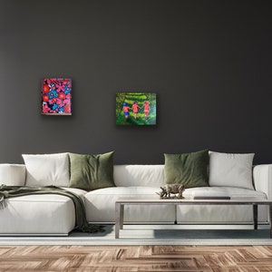 May include: Two paintings hanging on a dark gray wall above a white couch. The painting on the left features a colorful floral pattern. The painting on the right shows three people walking in a green field.