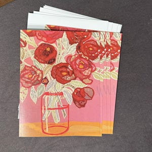 May include: A stack of greeting cards featuring a floral design. The cards showcase a bouquet of red roses in a glass jar against a pink background. The artwork has a collage-like appearance with various textures and patterns.