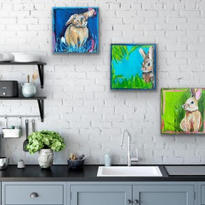 May include: Three square paintings of rabbits on a white brick wall. The paintings feature rabbits in a field of grass, with blue, green, and pink backgrounds. The rabbits are painted in shades of brown and white.