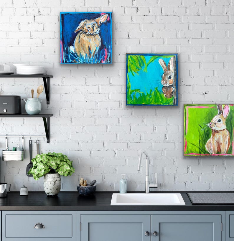 May include: Three square paintings of rabbits on a white brick wall. The paintings feature rabbits in a field of grass, with blue, green, and pink backgrounds. The rabbits are painted in shades of brown and white.