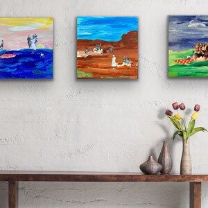 May include: Three framed paintings on a white wall, with a wooden table and a vase of flowers in front of them. The paintings are abstract and feature a variety of colors, including blue, green, red, and yellow.