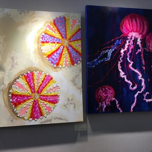 May include: Two paintings on canvas, one depicting three pink jellyfish with long tentacles on a dark blue background, the other depicting two colorful sea urchins on a light beige background.