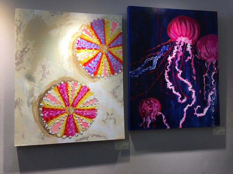 May include: Two paintings on canvas, one depicting three pink jellyfish with long tentacles on a dark blue background, the other depicting two colorful sea urchins on a light beige background.