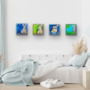 Peut inclure: Quatre peintures carr&eacute;es de lapins dans diverses poses. Chaque peinture pr&eacute;sente un lapin diff&eacute;rent sur un fond color&eacute;. Les peintures sont expos&eacute;es au-dessus d'un lit avec une couverture bleu clair.