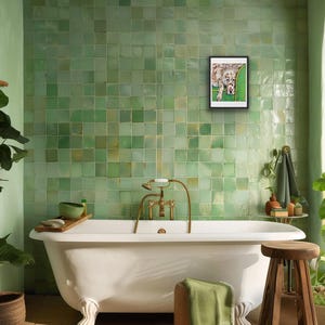May include: A framed rectangular wall art print featuring a spotted dog resting on a green sofa, displayed on a wall covered in square light green tiles above a white clawfoot bathtub with brass fixtures and a wooden stool with a green towel.