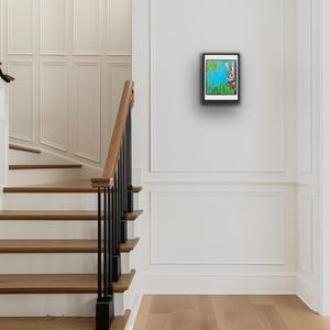 Op de afbeelding: Een ingelijst kunstwerk met een konijn dat achter groene bladeren vandaan gluurt tegen een blauwe achtergrond. Het kunstwerk hangt aan een witte muur in een lichte hal met houten trappen en een zwarte leuning.