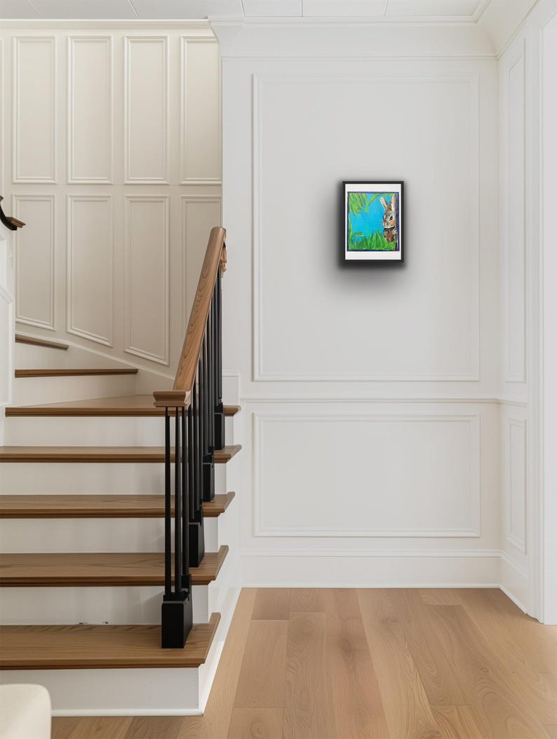 Op de afbeelding: Een ingelijst kunstwerk met een konijn dat achter groene bladeren vandaan gluurt tegen een blauwe achtergrond. Het kunstwerk hangt aan een witte muur in een lichte hal met houten trappen en een zwarte leuning.