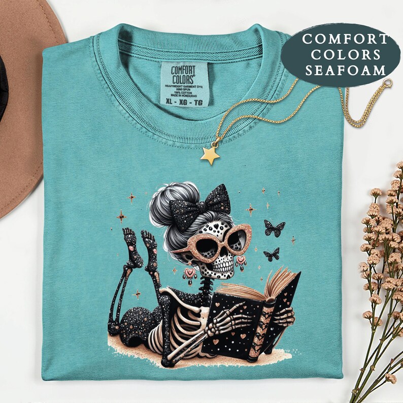 May include: A seafoam green Comfort Colors t-shirt featuring a graphic of a skeleton with a hair bow, glittery glasses, and earrings, lying down while reading an open book. The design includes small butterflies and stars, with a gold star necklace.