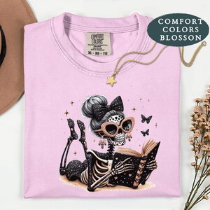 May include: A light pink t-shirt featuring a graphic of a skeleton with a messy bun, a black bow, and glittery rose gold sunglasses, lying down and reading a book. The shirt has a Comfort Colors label at the neck and a star-shaped pendant necklace.