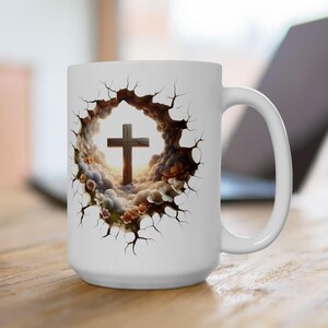 May include: White ceramic mug featuring a detailed graphic of a wooden cross set against a backdrop of clouds and flowers. The design is framed by a cracked, textured border. The mug has a large handle and is set on a wooden surface.