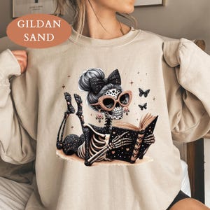 May include: A sand-colored crewneck sweatshirt featuring a graphic of a skeleton lying down and reading a book. The skeleton has a high bun with a black bow, rose gold glitter sunglasses, and dangling earrings. Small butterflies and stars surround the figure.