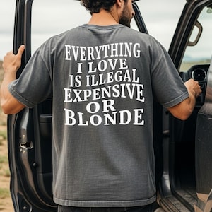May include: Dark gray t-shirt with the text "EVERYTHING I LOVE IS ILLEGAL EXPENSIVE OR BLONDE" in white block letters. The shirt is a short-sleeved crew neck style. The person is standing next to a vehicle.