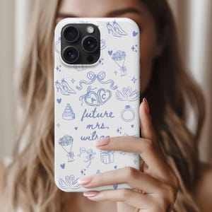 May include: White phone case with a blue illustrated design featuring wedding-themed icons like shoes, bouquets, rings, and swans. The text "future mrs." is also included in the design. The case is held in front of a person.