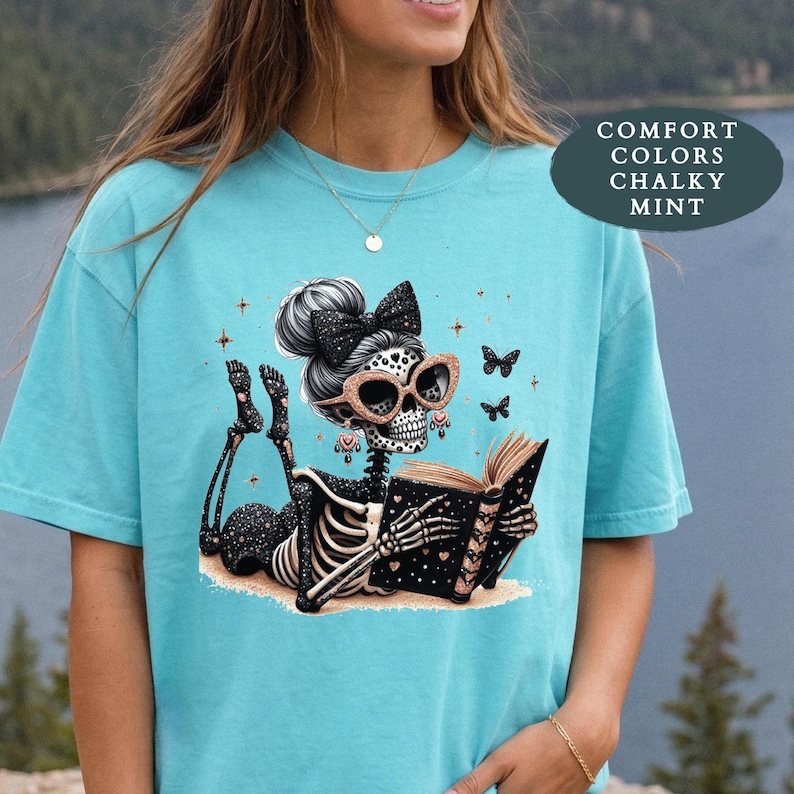 May include: A mint green t-shirt featuring a graphic of a skeleton with a high bun and a black bow, wearing glittery glasses and reading a black book with gold hearts. The design includes small stars and butterflies, with text reading COMFORT COLORS CHALKY MINT.