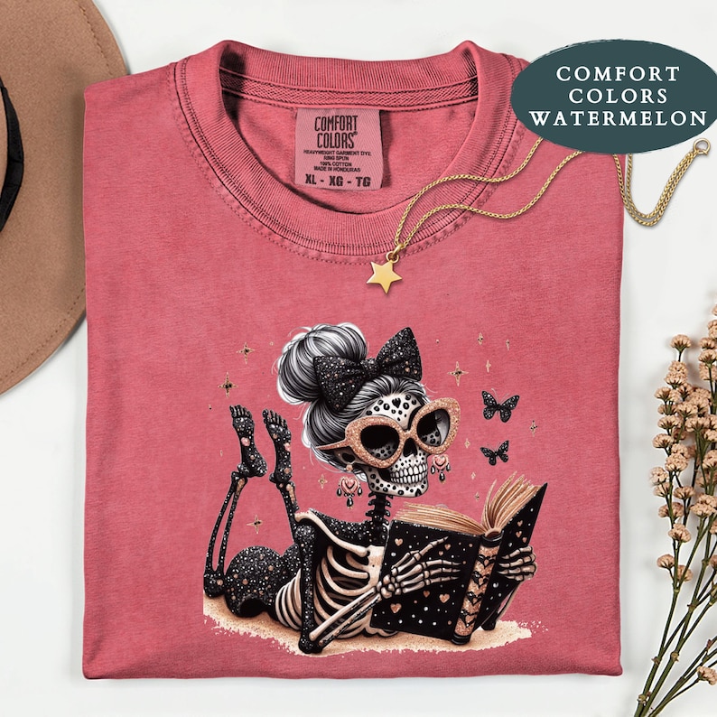 May include: A watermelon-colored t-shirt featuring a graphic of a skeleton with a high bun, a black bow, and glittery cat-eye glasses, lying down and reading a book. The shirt has a Comfort Colors label and is paired with a gold star necklace.