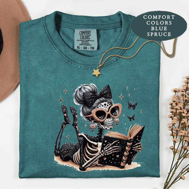 May include: A blue spruce Comfort Colors t-shirt featuring a graphic of a skeleton with a high bun, a black bow, and glittery glasses reading an open book. The skeleton is decorated with small hearts and stars. A gold star necklace rests on the shirt.