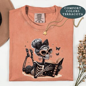 May include: A terracotta-colored t-shirt featuring a graphic of a skeleton with a hair bow and glasses reading a book. The skeleton has star patterns on its body. The shirt has a Comfort Colors label at the neck and a gold star necklace.