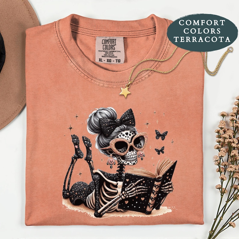 May include: A terracotta-colored t-shirt featuring a graphic of a skeleton with a hair bow and glasses reading a book. The skeleton has star patterns on its body. The shirt has a Comfort Colors label at the neck and a gold star necklace.