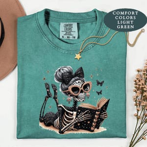 May include: A light green t-shirt featuring a graphic of a skeleton with a high bun, a black bow, and glittery rose gold sunglasses, lying down and reading a book. The skeleton is surrounded by small butterflies and stars. The shirt has a Comfort Colors label.