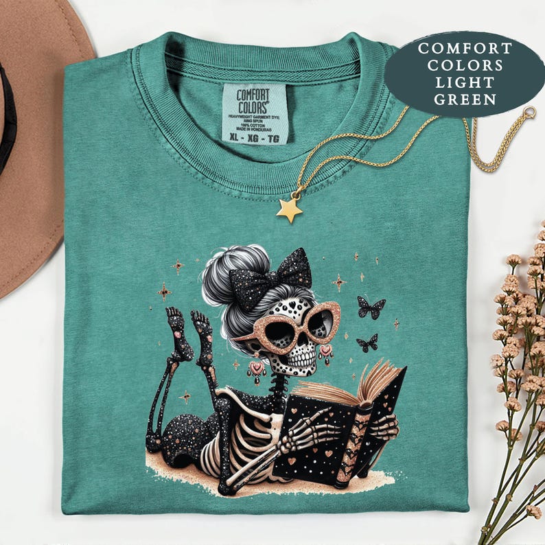 May include: A light green t-shirt featuring a graphic of a skeleton with a high bun, a black bow, and glittery rose gold sunglasses, lying down and reading a book. The skeleton is surrounded by small butterflies and stars. The shirt has a Comfort Colors label.