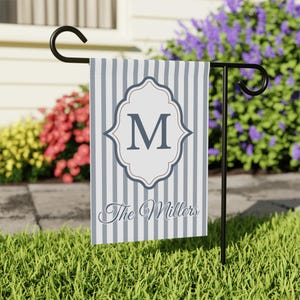 May include: A garden flag with vertical gray and white stripes. The flag features a white, ornate frame with the letter "M" in the center. Below the frame, the text "The Millers" is written in cursive. The flag is displayed on a black metal stand.