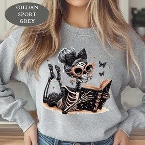 May include: A Gildan Sport Grey sweatshirt featuring a graphic of a skeleton with a high bun, a black bow, and rose gold glasses reading a book. The skeleton has black and rose gold accents, including heart patterns and small butterflies.