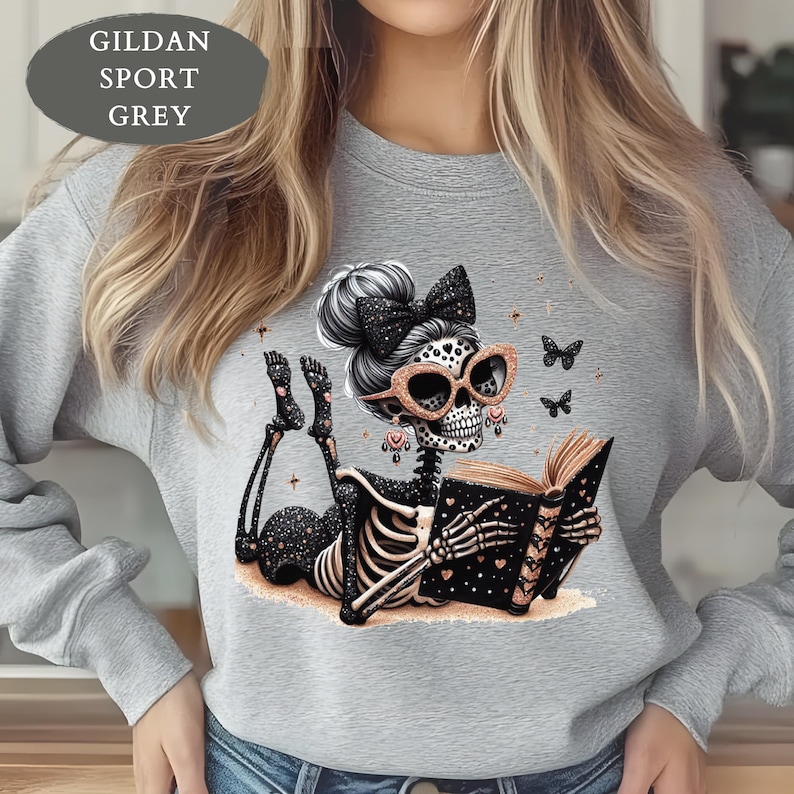 May include: A Gildan Sport Grey sweatshirt featuring a graphic of a skeleton with a high bun, a black bow, and rose gold glasses reading a book. The skeleton has black and rose gold accents, including heart patterns and small butterflies.