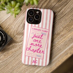 Puede incluir: Funda de tel&eacute;fono a rayas rosas y blancas. Presenta un dise&ntilde;o de tarjeta de biblioteca rosa con el texto "just one more chapter". La funda tiene un recorte para c&aacute;mara de triple lente y las iniciales "AC" en la parte inferior.