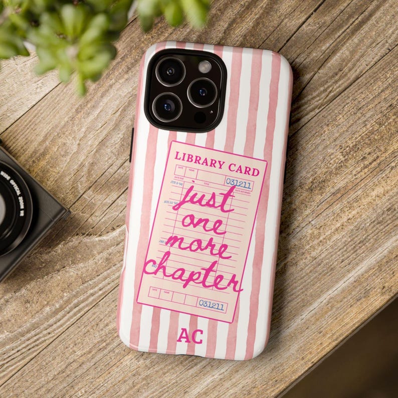 Puede incluir: Funda de tel&eacute;fono a rayas rosas y blancas. Presenta un dise&ntilde;o de tarjeta de biblioteca rosa con el texto "just one more chapter". La funda tiene un recorte para c&aacute;mara de triple lente y las iniciales "AC" en la parte inferior.