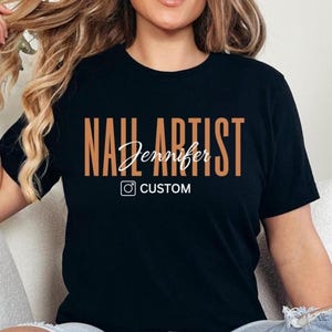 May include: A black short-sleeved t-shirt featuring the text NAIL ARTIST in large tan letters, the name Jennifer in white script, and a white Instagram icon followed by the word CUSTOM printed on the front center of the garment.