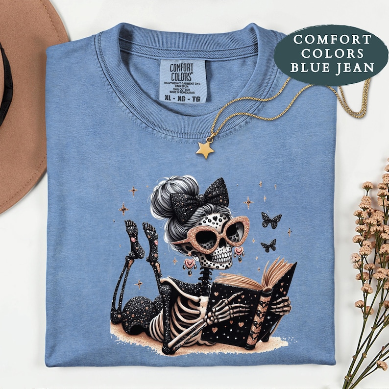 May include: A blue jean colored t-shirt with a Comfort Colors label at the neck. The graphic features a skeleton with a messy bun, a black bow, and rose gold glasses, lying down while reading a black book with gold hearts and stars.