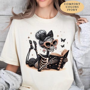 May include: An ivory t-shirt featuring a graphic of a skeleton with a black bow in its hair, wearing glittery cat-eye glasses, and reading a book. The skeleton is surrounded by small stars and butterflies. Text on the shirt reads COMFORT COLORS IVORY.