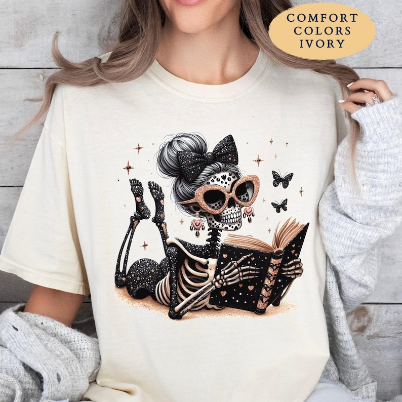 May include: An ivory t-shirt featuring a graphic of a skeleton with a black bow in its hair, wearing glittery cat-eye glasses, and reading a book. The skeleton is surrounded by small stars and butterflies. Text on the shirt reads COMFORT COLORS IVORY.