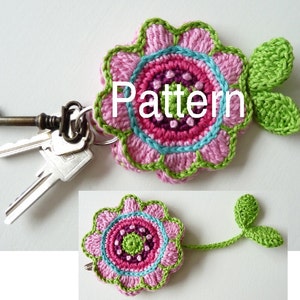 Puede incluir: Un llavero de flor de ganchillo hecho a mano en rosa, verde y azul. La flor tiene un diseño detallado con un círculo central y bordes en forma de pétalos. La palabra "Pattern" está escrita en la flor. Un juego de llaves está unido al llavero.