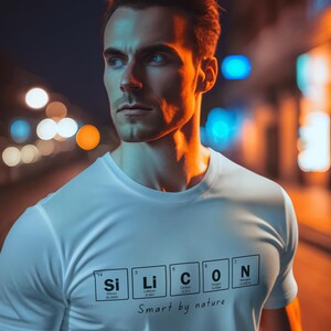 May include: White t-shirt with the word "SiLiCON" in a periodic table style, with the text "Smart by nature" underneath. The image is of a person wearing the shirt, with a blurred background of city lights.