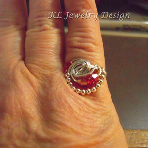 May include: A handmade ring with a heart-shaped design. The ring features a large, faceted red crystal at the center, surrounded by a silver wire swirl and a border of small silver beads. The text "KL Jewelry Design" is visible.