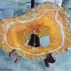 May include: A decorative agate slice with a natural, organic shape. The slice features warm tones of orange and yellow, with white and clear crystalline edges. It is displayed on a wooden stand, showcasing its unique, artistic design.