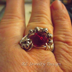 Op de afbeelding: Een zilveren ring met een rode edelsteen gezet in een draadgewikkeld ontwerp. De ring zit aan een vinger.