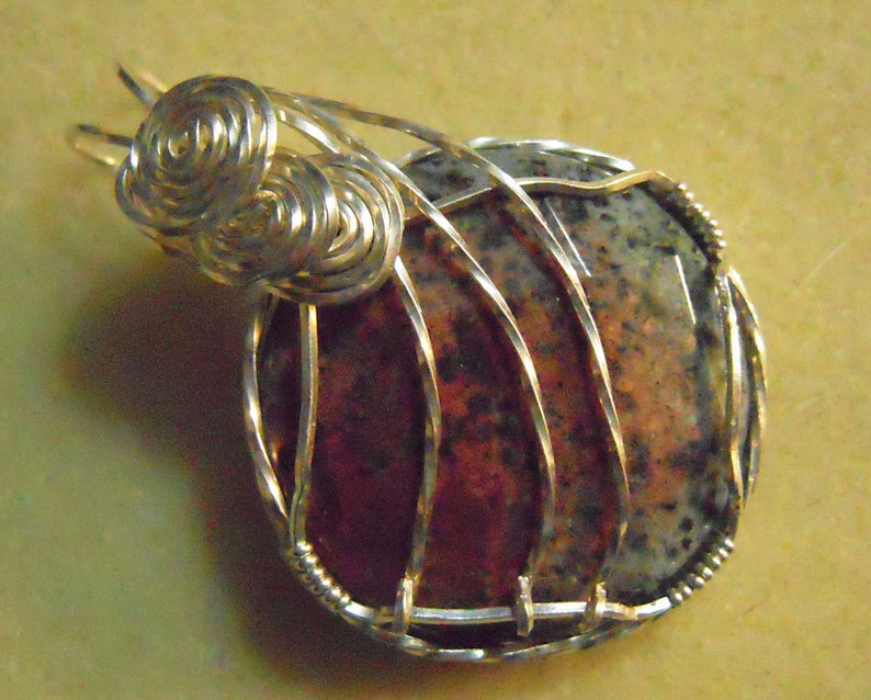 May include: A silver wire wrapped pendant featuring a round, multi-colored stone with a mottled pattern. The wire is twisted into a decorative design at the top of the pendant.