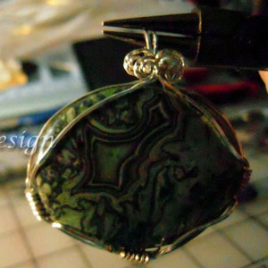 May include: A green and brown agate stone pendant wrapped in silver wire. The stone has a swirling pattern and is set in a silver wire frame.