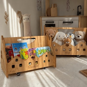 May include: Two wooden toy storage carts with books and stuffed animals. The carts are light brown with a house-shaped design and wheels. One cart has the name "Martin" and the other has the name "Mia" on it.