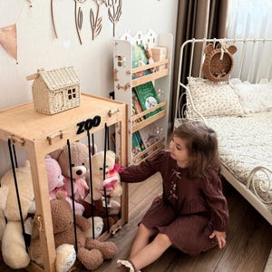 Op de afbeelding: Een kinderkamersc&egrave;ne met een houten speelgoedzoo vol teddyberen. Een kleine huisvormige mand staat bovenop de zoo. Een witte boekenplank en een bed met bloemenkussens zijn ook zichtbaar. Het woord "ZOO" staat op de voorkant van het speelgoed.