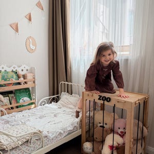 Op de afbeelding: Kinderslaapkamer met een houten speelgoedopbergdoos met het opschrift "ZOO", gevuld met teddyberen. Een wit bed met een bloemenpatroon dekbed en een boekenplank met boeken zijn ook zichtbaar. De kamer wordt verlicht door natuurlijk licht.