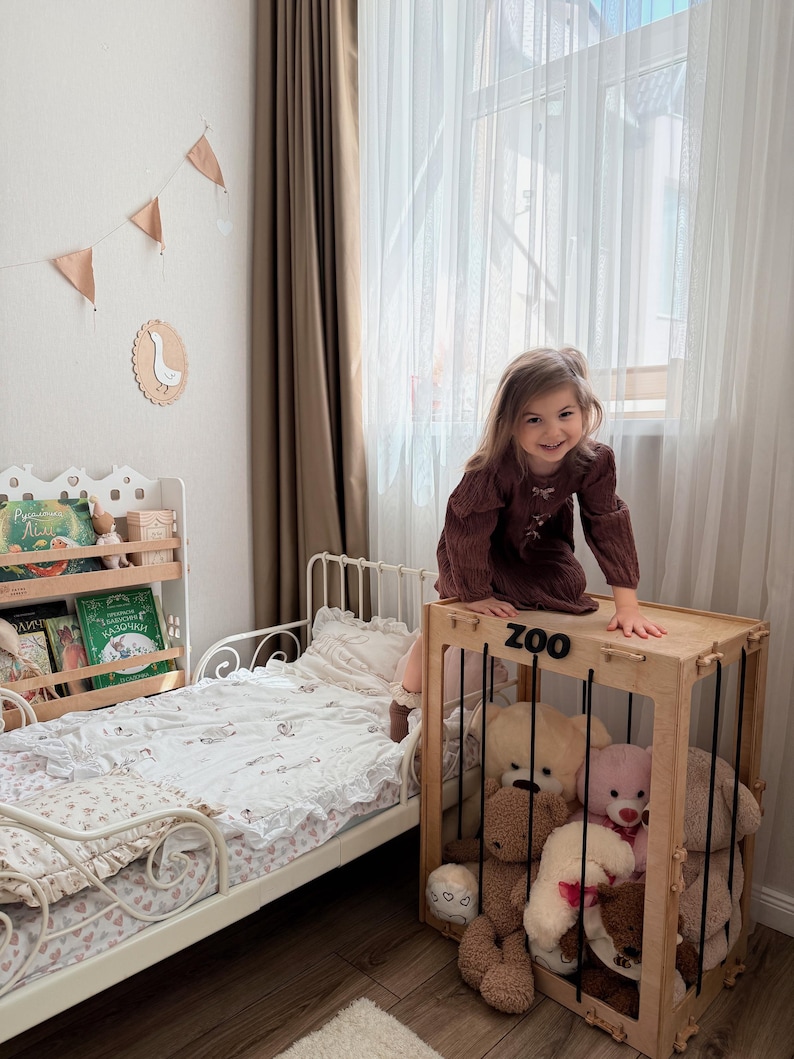Op de afbeelding: Kinderslaapkamer met een houten speelgoedopbergdoos met het opschrift "ZOO", gevuld met teddyberen. Een wit bed met een bloemenpatroon dekbed en een boekenplank met boeken zijn ook zichtbaar. De kamer wordt verlicht door natuurlijk licht.