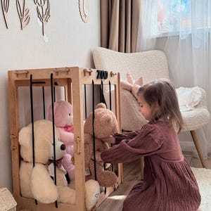 Op de afbeelding: Een houten speelgoed zoo-omheining met zwarte spijlen, met verschillende knuffelberen in verschillende kleuren. Het woord "ZOO" is zichtbaar aan de bovenkant. Een kind in een bordeauxrode jurk interacteert met het speelgoed.