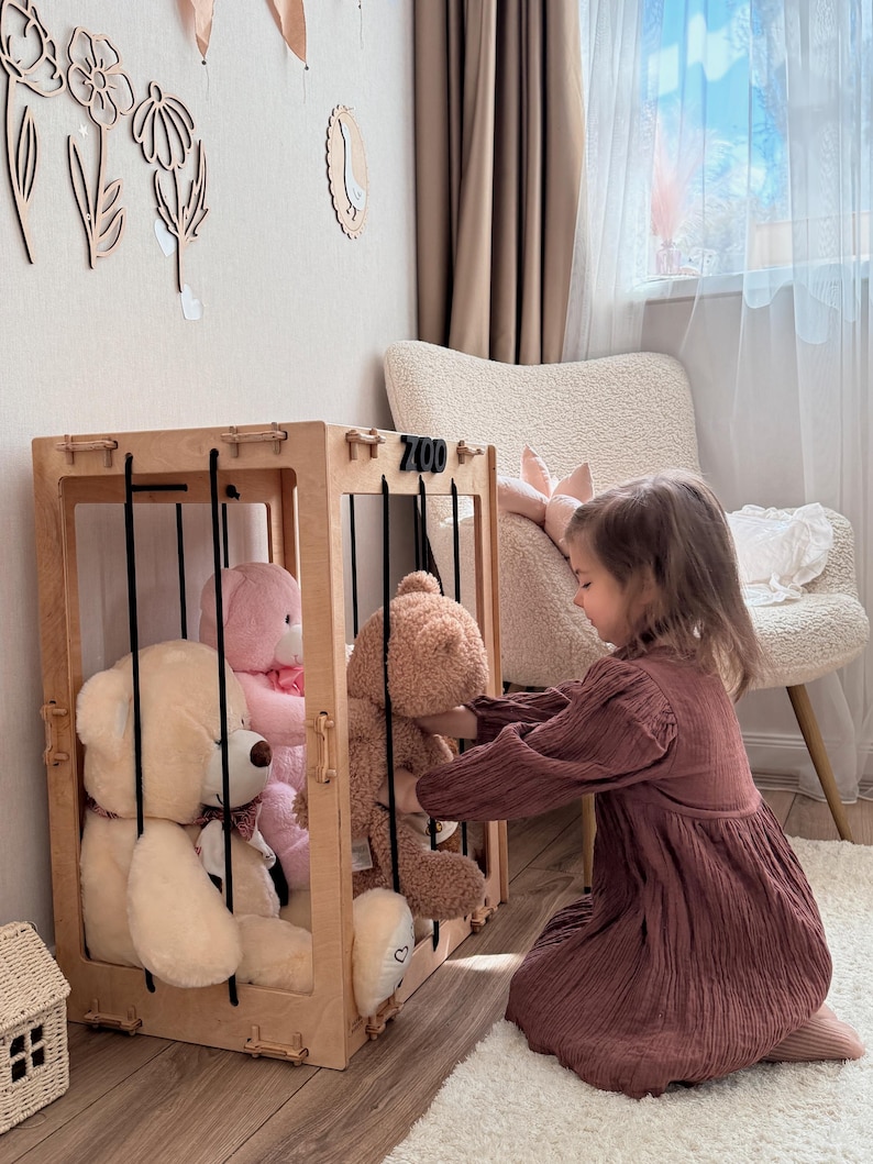 Op de afbeelding: Een houten speelgoed zoo-omheining met zwarte spijlen, met verschillende knuffelberen in verschillende kleuren. Het woord "ZOO" is zichtbaar aan de bovenkant. Een kind in een bordeauxrode jurk interacteert met het speelgoed.