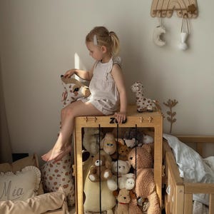 Op de afbeelding: Een houten speelgoedkist vol knuffels, met een kind dat erop zit. De kist is rechthoekig met een rooster aan de voorkant. Een houten regenboog wanddecoratie en een klein houten bed zijn ook zichtbaar.
