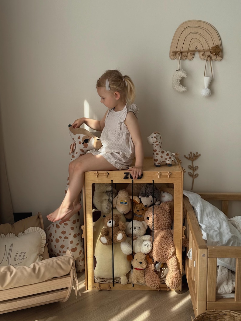 Op de afbeelding: Een houten speelgoedkist vol knuffels, met een kind dat erop zit. De kist is rechthoekig met een rooster aan de voorkant. Een houten regenboog wanddecoratie en een klein houten bed zijn ook zichtbaar.