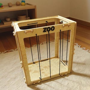 Op de afbeelding: Een houten speelgoed dierentuin kooi met zwarte verticale staven en het woord "ZOO" in zwarte letters. De kooi is gemaakt van lichtgekleurd hout en staat op een beige tapijt. Andere houten speelgoed is zichtbaar op de achtergrond.