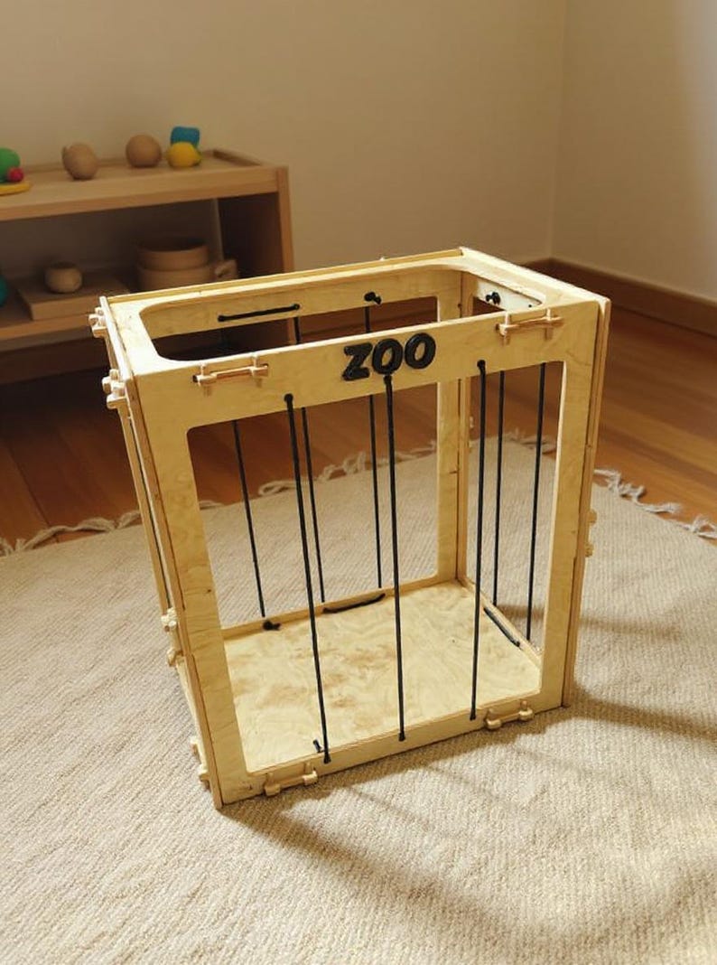 Op de afbeelding: Een houten speelgoed dierentuin kooi met zwarte verticale staven en het woord "ZOO" in zwarte letters. De kooi is gemaakt van lichtgekleurd hout en staat op een beige tapijt. Andere houten speelgoed is zichtbaar op de achtergrond.